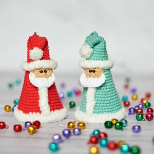 K&ouml;nnte beinhalten: Zwei geh&auml;kelte Weihnachtsmann-Ornamente, eines rot und eines blaugr&uuml;n, mit wei&szlig;em Besatz. Die Ornamente liegen auf einer wei&szlig;en Oberfl&auml;che mit bunten Dekorationskugeln, die um sie herum verstreut sind.
