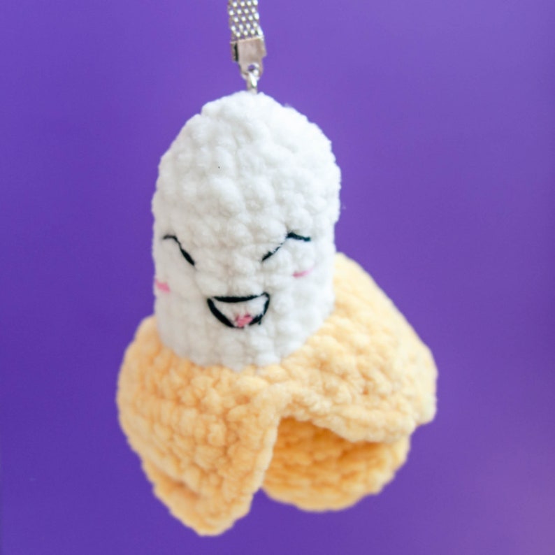 May include: A crocheted banana keychain with a smiling face. The banana is yellow and white with a silver chain.