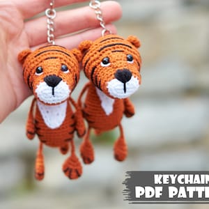 Peut inclure: Deux patrons de porte-clés en crochet en forme de tigre. Les tigres sont orange et noir avec des visages blancs. Le porte-clés est attaché à une chaîne argentée.