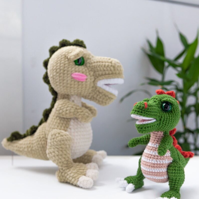 May include: Two crocheted dinosaur toys. The larger dinosaur is green and beige with a pink blush on its cheeks. The smaller dinosaur is green with a pink stripe around its middle and red accents.