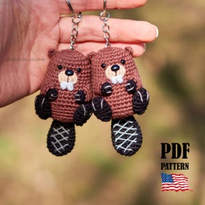 May include: Two handmade crochet beaver keychains. The beavers are brown with black paws and tails, white teeth, and small black eyes. Each keychain is attached to a silver chain. The image also includes the text "PDF PATTERN" and an American flag.