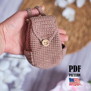Puede incluir: Una pequeña mochila de ganchillo en color rosa empolvado con cierre de solapa asegurado por un botón de madera redondo grabado con el texto "handmade". El artículo incluye dos correas superiores y bolsillos laterales con tejido denso.