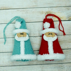 K&ouml;nnte beinhalten: Zwei geh&auml;kelte Weihnachtsmann-Anh&auml;nger, einer in Rot und einer in Blau, mit wei&szlig;em Besatz und schwarzen Augen. Die Anh&auml;nger h&auml;ngen an einem wei&szlig;en Holzgrund.