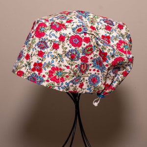 May include: A floral scrub cap featuring a white base with a colourful pattern of pink, blue, and green flowers and foliage. The cap is gathered at the back and has an adjustable toggle.