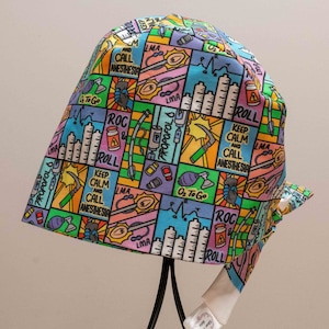May include: A colourful surgical scrub hat with a cartoon design featuring medical equipment and the text "Keep Calm and Call Anesthesia".