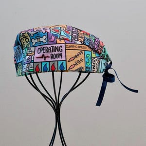 May include: A scrub cap featuring a colourful comic book style print with various medical illustrations and text including "OPERATING ROOM" and "CLAMP, CLAMP, CUT, TIE". The cap has a dark blue ribbon tie at the back for an adjustable fit.