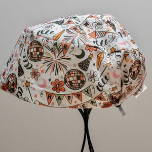 May include: A white fabric hat with a celebratory print. The design includes disco balls, fireworks, flowers, and the word "HAPPY" on a banner. The accents are orange, pink, and black, creating a festive theme.