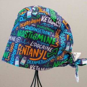 May include: A navy blue surgical scrub cap featuring a vibrant pattern of medical drug names and illustrations. The text includes "Fentanyl", "Ketamine", and "Neostigmine" in various colours. The cap has a white tie closure.