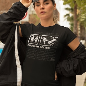 Può includere: Maglietta nera con una grafica bianca di due figure stilizzate. La prima figura stilizzata mostra un uomo e una donna che litigano. La seconda figura stilizzata mostra l'uomo che scappa. Il testo "PROBLEMA RISOLTO" è sotto la grafica.