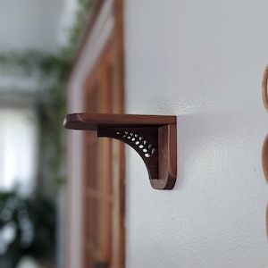 Pode incluir: Um suporte de prateleira de madeira castanho escuro com um design de recorte decorativo. O suporte est&aacute; montado numa parede branca.