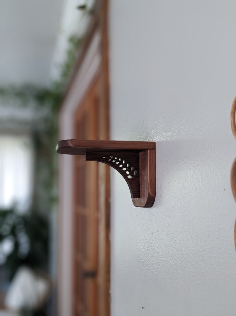 Pode incluir: Um suporte de prateleira de madeira castanho escuro com um design de recorte decorativo. O suporte est&aacute; montado numa parede branca.