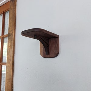 Pode incluir: Uma prateleira de parede de madeira castanho escuro com a parte superior curva e um design talhado decorativo. A prateleira est&aacute; montada numa parede branca.