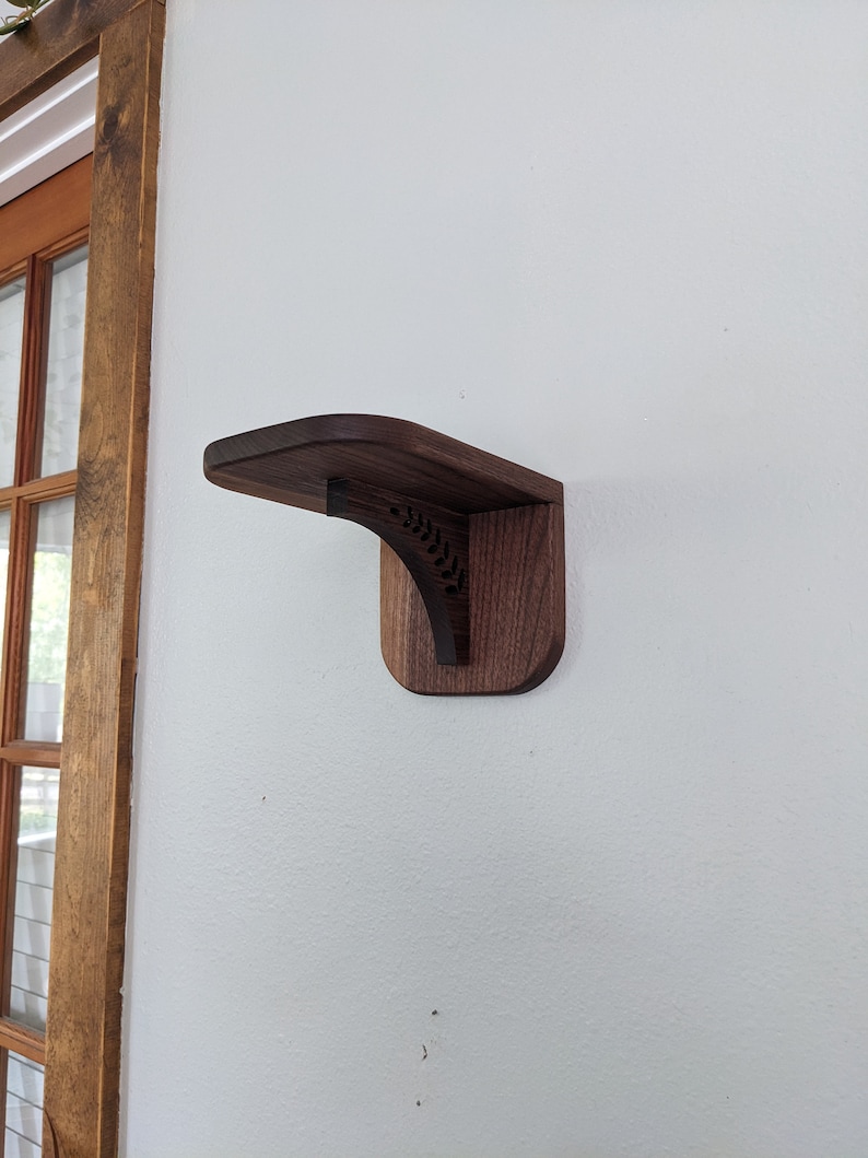 Pode incluir: Uma prateleira de parede de madeira castanho escuro com a parte superior curva e um design talhado decorativo. A prateleira est&aacute; montada numa parede branca.