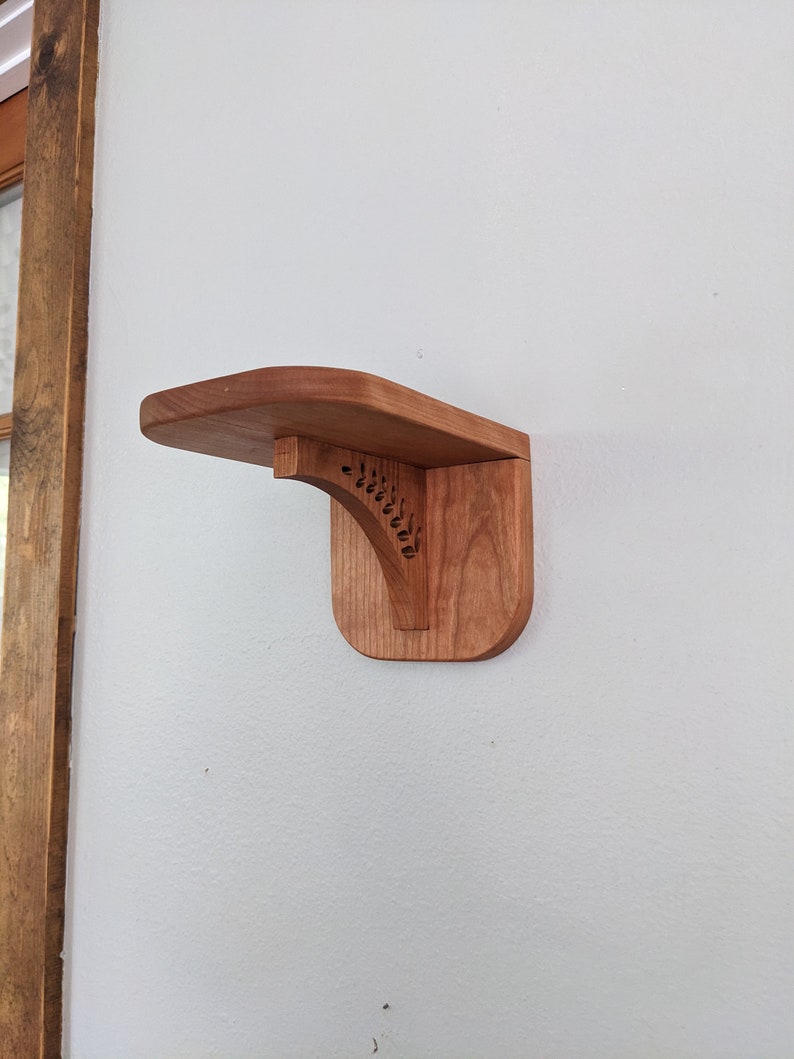 Pode incluir: Uma prateleira de madeira com um design decorativo esculpido no suporte. A prateleira est&aacute; montada em uma parede branca.