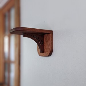 Pode incluir: Uma prateleira de madeira escura com um design de recorte decorativo est&aacute; montada em uma parede branca.