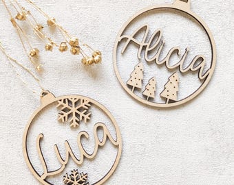 Könnte beinhalten: Zwei Weihnachtsbaumschmuck aus Holz. Ein Ornament trägt den Namen "Alicia" mit drei kleinen Weihnachtsbäumen. Das andere Ornament trägt den Namen "Luca" mit zwei Schneeflocken. Beide Ornamente sind rund und haben eine Schlaufe zum Aufhängen.
