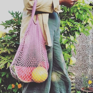 May include: A person carrying a pink mesh bag with a watermelon and a yellow fruit inside. The bag is hanging from their shoulder and they are wearing a plaid shirt and blue jeans.