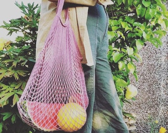 May include: A person carrying a pink mesh bag with a watermelon and a yellow fruit inside. The bag is hanging from their shoulder and they are wearing a plaid shirt and blue jeans.