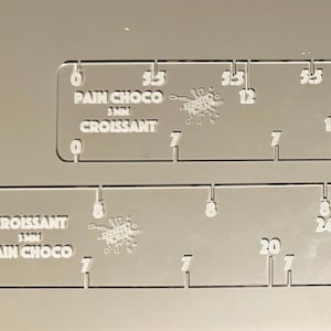 Può includere: Due righelli in acrilico trasparente con segni in millimetri. Il righello superiore ha il testo "PAIN CHOCO 3 MM CROISSANT" e il righello inferiore ha il testo "CROISSANT 3 MM PAIN CHOCO".