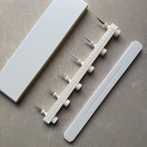 Peut inclure: Un ensemble d'outils de précision blancs pour l'artisanat. L'ensemble comprend une règle, une longue boîte rectangulaire et deux outils de mesure avec des lames tranchantes. Le texte "TAGLIOPERFETTO DESIGN FABRIZIOFIORANI 2025" est imprimé.
