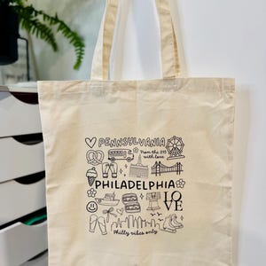 May include: Cream-coloured tote bag featuring black line art of Philadelphia landmarks and the words "PENNSYLVANIA" and "PHILADELPHIA". The bag includes the phrase "Philly vibes only" and a heart symbol.