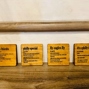 May include: Four square wooden coasters with phrases related to the Philadelphia Eagles football team. Each coaster has a different phrase in black text: "go birds", "philly special", "fly eagles fly", and "it's a philly thing".