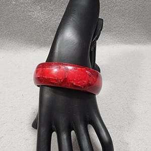 Puede incluir: Un brazalete rígido de color rojo translúcido que presenta un patrón interno profundo y multicapa con diversas tonalidades de carmesí y escarlata. El accesorio circular tiene una superficie lisa y pulida con un perfil grueso y redondeado.