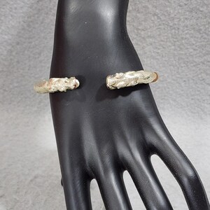 May include: A gold-toned cuff bracelet featuring two sculpted dragon heads facing each other at the opening. The metal surface displays intricate, textured detailing across the length of the band, showcasing a polished finish with subtle highlights.