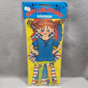 Può includere: Un set di bambola di carta Pippi Langstrumpf nella confezione originale. Il personaggio ha capelli rossi intrecciati, lentiggini e occhi azzurri. La bambola indossa una maglietta blu, pantaloncini gialli e calze spaiate verdi e gialle.