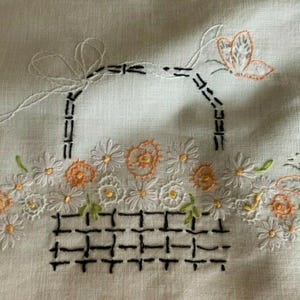 May include: A white fabric piece featuring hand embroidery of a black basket filled with white daisies and orange flowers. The design includes two orange butterflies, one on each side, and a black handle with a white bow tied at the top left corner.