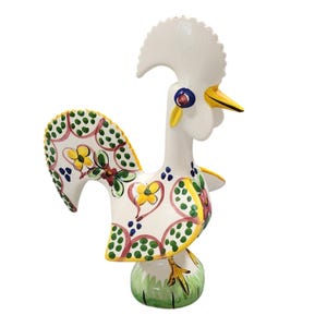 Puede incluir: Figura de gallo de cerámica con cuerpo blanco, pico amarillo y gran cresta blanca. Las alas y la cola del gallo están decoradas con motivos florales en amarillo, verde y rojo. La base es verde.