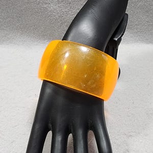 May include: A wide, translucent orange bangle bracelet with a smooth, polished surface. The bracelet features a simple, continuous circular design with a high-gloss finish that catches the light, showcasing its vibrant, semi-transparent color.