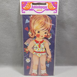 Può includere: Un set di bambole di carta vintage nella confezione originale etichettata Ankleidepuppe siebert. La bambola ha capelli biondi, guance rosate e un costume da bagno a fiori. Lo sfondo è un cartoncino viola scuro con illustrazioni di cuori e fiori.