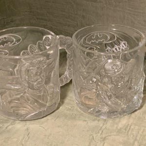 May include: Two clear glass mugs with handles. Each mug features embossed images of Batman characters and the Batman logo. The mugs are sitting on a light green surface.