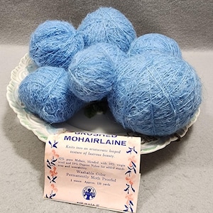 Op de afbeelding: Zes bollen lichtblauw mohairgaren liggen op een decoratief bord. Een vintage label leest "Brushed Mohairlaine" en geeft de samenstelling van het garen weer, waaronder 62% mohair, 19% scheerwol en 19% Dupont Nylon. Het label vermeldt ook dat het garen wasbaar en motbestendig is.
