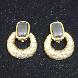 May include: A pair of gold-tone earrings featuring a rectangular black stone set at the top, connected to a circular hoop with a textured, scale-like pattern. The earrings have a polished finish and a classic design with a secure post-style backing.