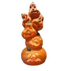 Può includere: Una pila verticale di cinque zucche di Halloween arancioni con volti intagliati. Ogni zucca presenta ritagli per occhi, naso e bocca, con una fonte di luce interna che emette un caldo bagliore arancione attraverso le aperture.