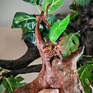 May include: A brown, gnarled, tree-like creature with red eyes and a toothy grin. The creature is made of clay and is surrounded by green leaves.
