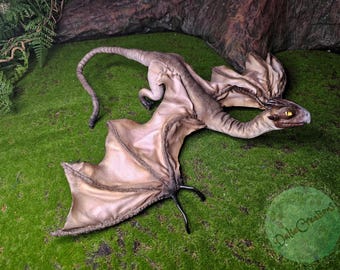 Puede incluir: Escultura detallada de un pequeño dragón con alas, cuerpo y cabeza de color marrón y beige, con ojos amarillos. El dragón está posado sobre musgo verde, con un fondo oscuro. Las alas del dragón están extendidas y su cola está curvada.