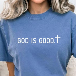 May include: A blue short-sleeved t-shirt with the text "GOD IS GOOD" in white, accompanied by a white cross symbol. The shirt is made of a soft material and is suitable for casual wear.