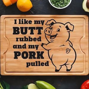 May include: A rectangular wooden cutting board with a carved handle. The board features the text "I like my BUTT rubbed and my PORK pulled" and a cartoon pig illustration. The board is surrounded by fresh produce.