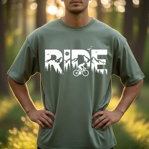 Op de afbeelding: Olijfgroen T-shirt met een witte opdruk van 'RIDE' met een berg- en fietsontwerp. Het shirt met korte mouwen heeft een relaxte pasvorm. De persoon draagt het shirt buiten.