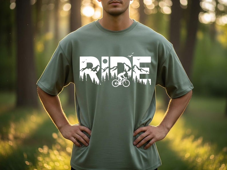 Op de afbeelding: Olijfgroen T-shirt met een witte opdruk van 'RIDE' met een berg- en fietsontwerp. Het shirt met korte mouwen heeft een relaxte pasvorm. De persoon draagt het shirt buiten.