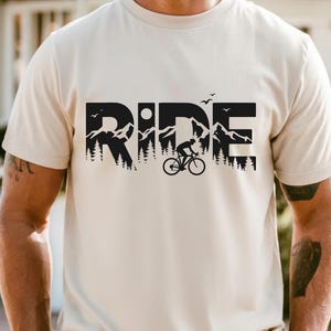 Op de afbeelding: Een lichtbeige t-shirt met een zwart grafisch ontwerp. Het woord "RIDE" wordt weergegeven in grote, vette letters, met een berg- en boslandschap verwerkt in de letters. Een fietser op een fiets is ook te zien.