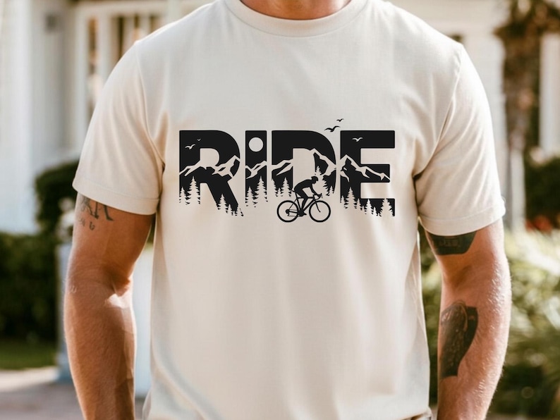 Op de afbeelding: Een lichtbeige t-shirt met een zwart grafisch ontwerp. Het woord "RIDE" wordt weergegeven in grote, vette letters, met een berg- en boslandschap verwerkt in de letters. Een fietser op een fiets is ook te zien.