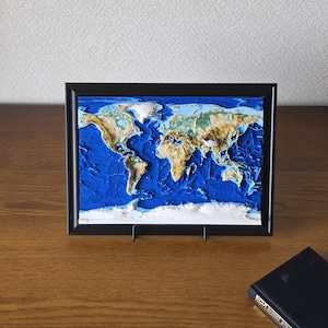 May include: A framed 3D relief map of the world featuring textured landmasses in shades of green, brown, and white, set against a deep blue ocean background. The continents are raised, showing topographical detail within a simple black rectangular frame.