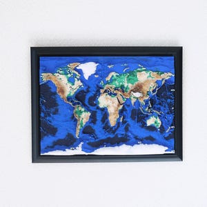 May include: A framed, three-dimensional world map. The map features a deep blue ocean, with landmasses in shades of brown, green, and white. The map is encased in a black frame and mounted on a white wall.