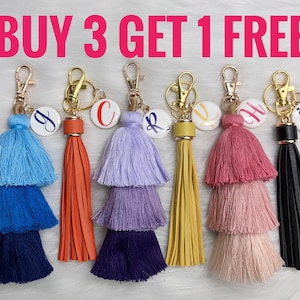 May include: Buy 3 get 1 free.  Set of six colorful tassel keychains with a round charm featuring a letter.  The charms are attached to a gold-colored metal ring.  The tassels are made of multi-colored yarn and are layered in three tiers.