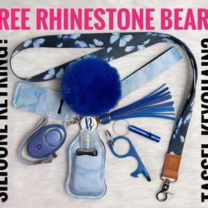 May include: A blue and black patterned lanyard with a blue pom-pom tassel, a blue silicone keychain with a hand sanitizer holder, a blue personal alarm, a blue and silver key ring, and a blue and silver tool. The lanyard has the text "FREE RHINESTONE BEAR?" and "TASSEL KEYCHAIN?" on it. The text "SILICONE KEYRING?" is on the left side of the image.