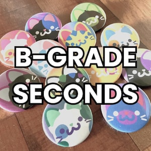 May include: A collection of colorful round buttons with cat face designs. The buttons feature various color schemes, including pink, blue, yellow, and green. The text "B-GRADE SECONDS" is displayed in a bold, white font across the image.